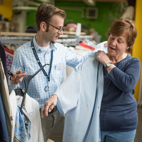Ein Aktiver der Tafel Jugend hilft einer Tafel-Kundin bei der Kleiderbörse, eine Jacke anzuziehen.